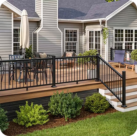 A backyard deck with black railings, patio furniture, and an umbrella sits attached to a gray house with large windows and a chimney—perfect inspiration for New Home Decks in San Antonio, Texas. Steps lead down to a landscaped garden with green shrubs.