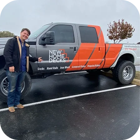 A person smiles and points at a large gray and orange New Home Decks pickup truck, advertising decks, hand rails, and iron work. The truck is parked outdoors on a cloudy day in San Antonio, Texas.