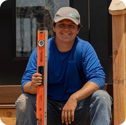 A smiling man in a blue shirt and gray cap sits on steps, holding an orange level tool. He is outside a new home deck in San Antonio, Texas, with a wooden post and a dark door visible behind him.
